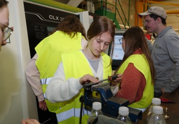 3 Schülerinnen stehen mit einem männlichen Lehrling vor einer CNC-Bearbeitungsmaschine, eine Schülerin feilt gerade ein Werkstück