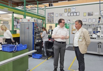 Michael Wölfl und Dr. Johannes Kopf befinden sich in der Lehrwerkstätte und unterhalten sich, im Hintergrund sieht man einen weiblichen Lehrling bei der Ausführung von elektrotechnischen Tätigkeiten