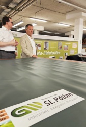 Michael Wölfl und Dr. Johannes Kopf stehen hinter einem Zuschnitttisch und sprechen miteinander, im Hintergrund sieht man eine „Werbetafel“ für Wohn-/Heimtextilien