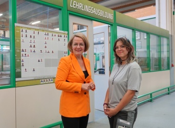 Eine ältere und eine junge Frau stehen lächelnd vor einer Tür. Darüber ein Schild 