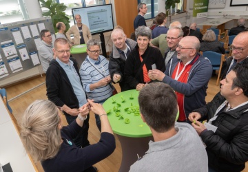 Eine Gruppe von Menschen steht um einen Runden Tisch, eine Gebärdendolmetscherin übersetzt