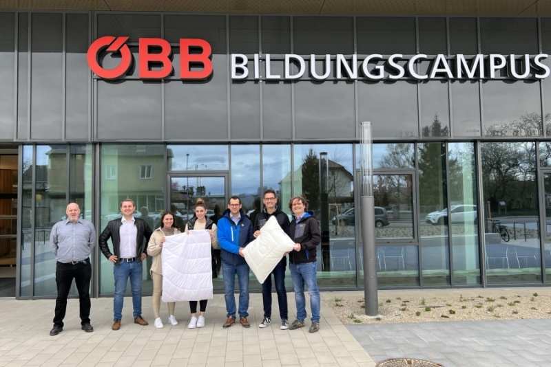 Gruppenfoto vor dem ÖBB Bildungscampus. Sie halten eine Decke und einen Polster in die Höhe.