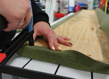 Nahaufnahme beim Polstern, eine Person befestigt gr&uuml;nen Stoff mit einem Drucklufttacker an einer Sitzunterlage aus Holz.