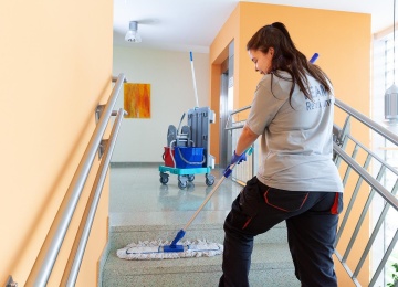 Reinigungstechnikerin wischt die Treppenstufen in einem hellen Stiegenhaus; neben ihr steht ein Reinigungswagen.