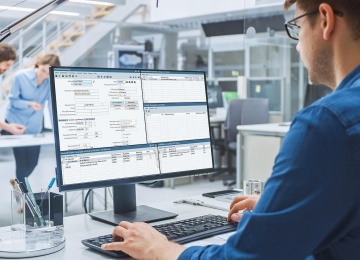 Ein Mitarbeiter sitzt an einem Arbeitsplatz und gibt Daten in eine Software am Computer ein; im Hintergrund arbeiten weitere Kollegen in einem modernen B&uuml;ro.