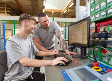 Lehrlingsausbilder arbeitet mit dem Lehrling an an einem Computerarbeitsplatz in einer Werkhalle.