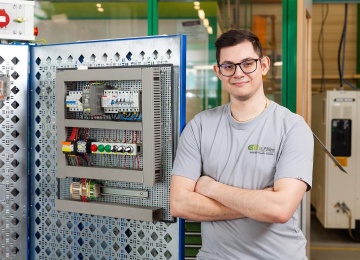 Lehrling steht neben einem ge&ouml;ffneten Schaltschrank mit elektrischen Komponenten.