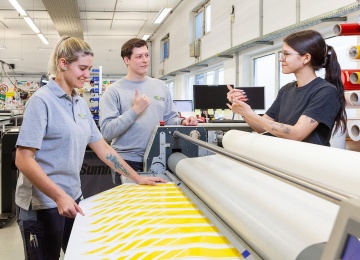 Drei Lehrlinge besprechen in Geb&auml;rden, an einer Druckmaschine die Verarbeitung einer bedruckten Folie.