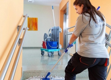 Reinigungstechnikerin wischt die Treppenstufen in einem hellen Stiegenhaus; neben ihr steht ein Reinigungswagen.