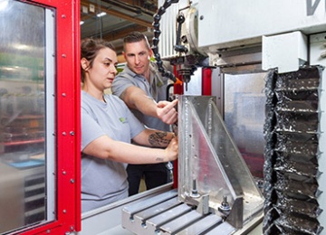 Junge Frau steht an Metallverarbeitungsmaschine, hinter ihr steht ein Ausbildner.