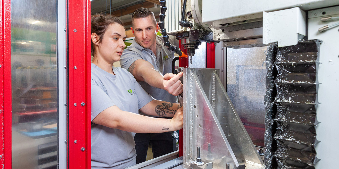 Junge Frau arbeitet an einer Metallbearbeitungsmaschine und erhält Instruktionen vom Mann neben ihr.