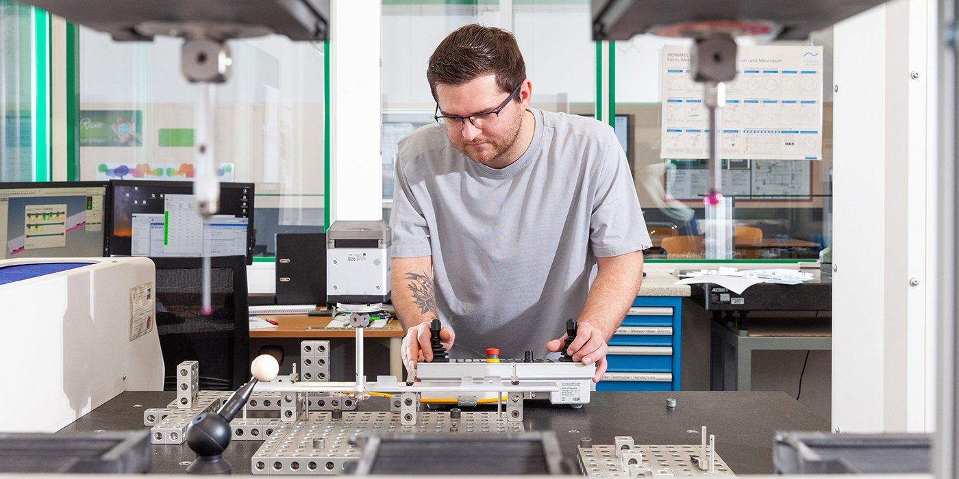 Junger Mann in blauen Shirt steht vor Maschine und steuert Metallteile.