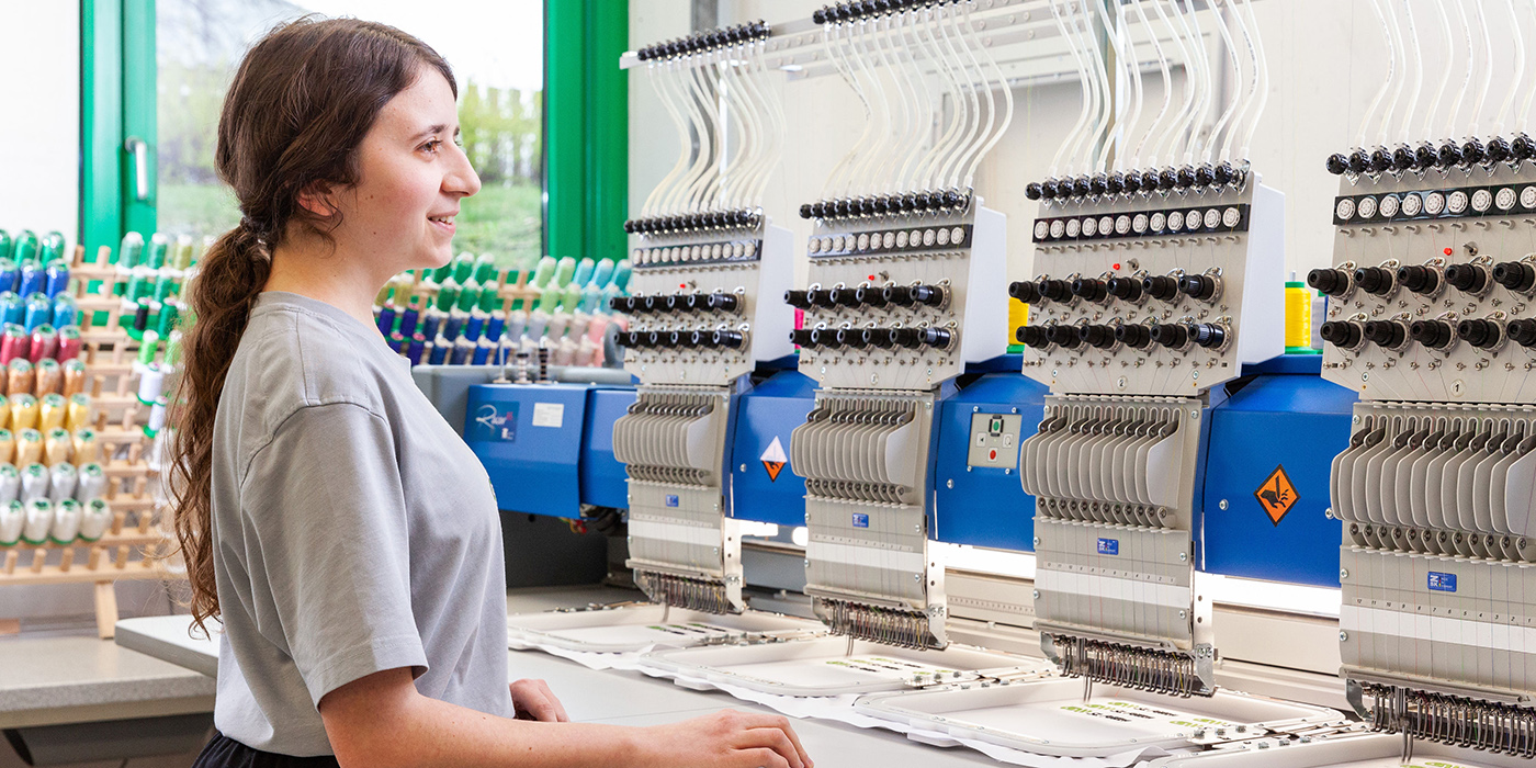 Mädchen steht lächelnd an einer großen Stickmaschine und überwacht den Arbeitsfortschritt.