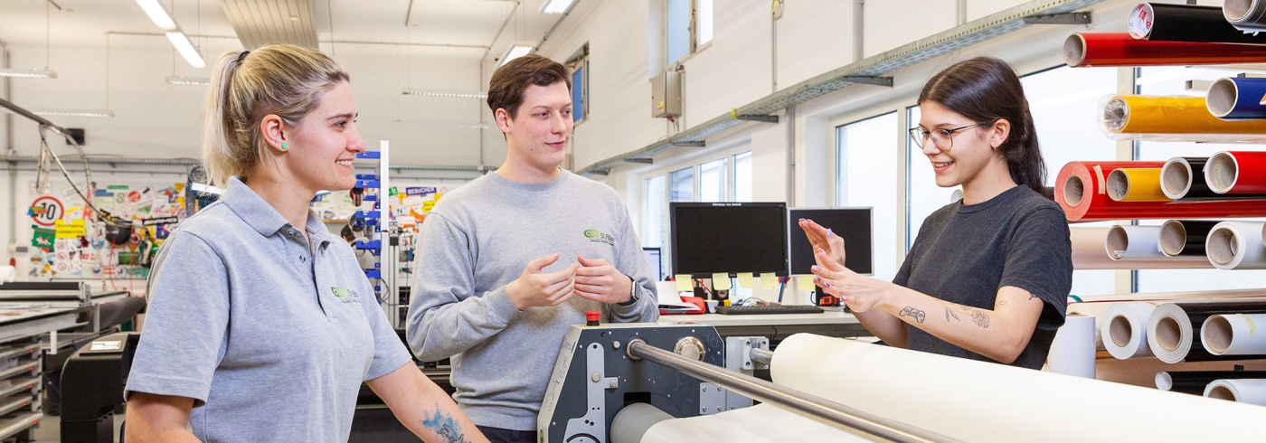 3 Junge Mitarbeiter stehen um eine Druckmaschine und unterhalten sich.