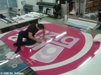 Ein Mann sitzt auf einem überdimensionalem großen rosa Werbeschild und beklebt Buchstaben