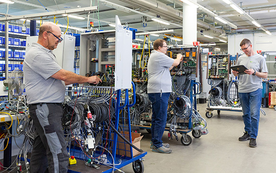 Drei Männer arbeiten lachend in einer Halle voller Kabel. Einer hält ein Klemmbrett.