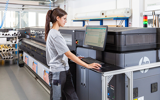 Ein Transparent auf der Druckmaschine. Davor eine junge Frau am Computer und Drucker.