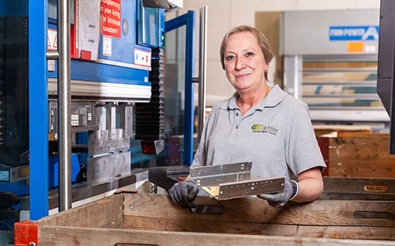 Eine Frau seht neben einer großen Maschine und hält ein Metall-Element in der Hand.