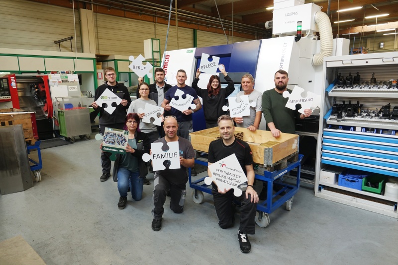 Gruppe von Mitarbeiter*innen stehen vor einer Produktionsmaschine und halten Puzzle-Teile mit unterschiedlichen Schlagworten in H&auml;nden, wie z.B. Vereinbarkeit von Beruf und Familie/Privatleben, Kommunikation, Pflege, Homeoffice, F&uuml;hrungsgrunds&auml;tze, Betriebsrat usw.