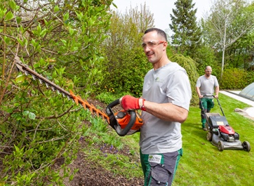 Ein Mann schneidet eine Hecke mit einer Heckenschere. Dahinter mäht ein anderer den Rasen.