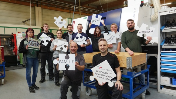 Gruppenfoto von Mitarbeiter*innen vor einer Produktionsmaschine mit Puzzleteilen in den H&auml;nden mit Themen wie Vereinbarkeit von Beruf und Familie/Privatleben, F&uuml;hrungsgrunds&auml;tze, Gemeinsam.Wir, Homeoffice, Pflege, Kommunikation, Betriebsrat,